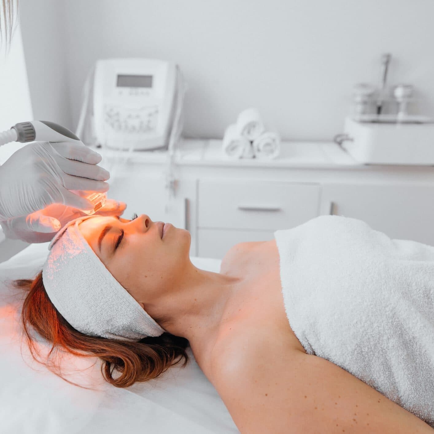 Woman receiving a red light therapy facial treatment from a gloved professional in a spa.