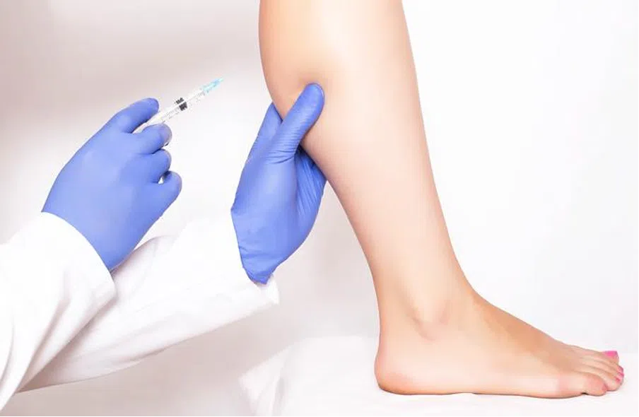 Medical professional in blue gloves holding a syringe to give an injection into a patient's calf.