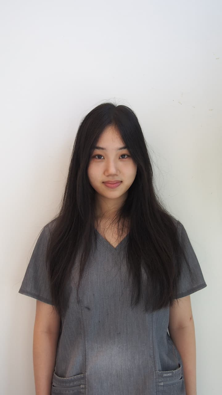 Young woman with long dark hair wearing a grey scrub top against a white wall.