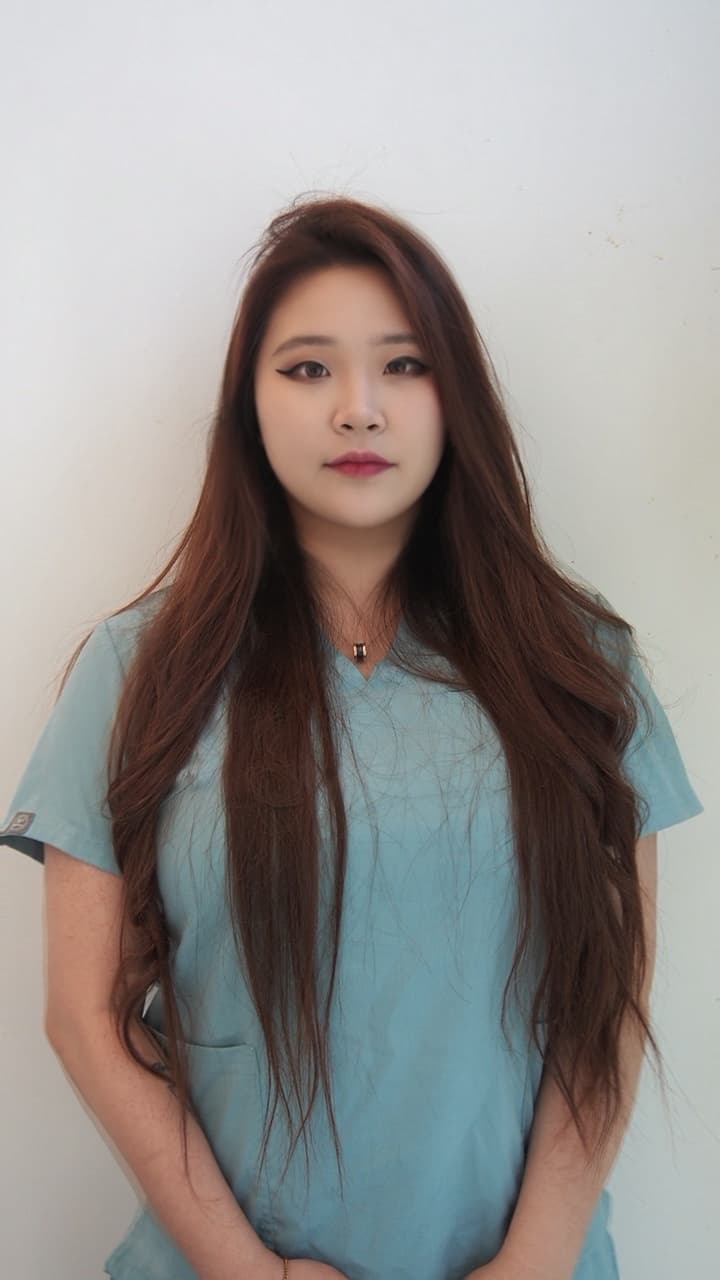 Young woman with long brown hair wearing light blue scrubs against a plain white wall.