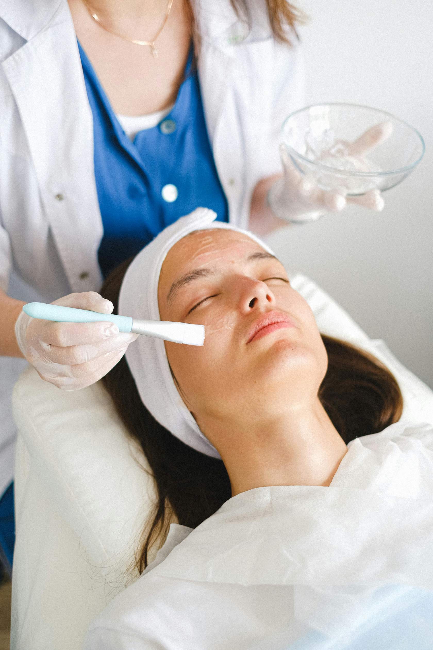 Esthetician applying clear gel mask to a woman's face with a brush during treatment.